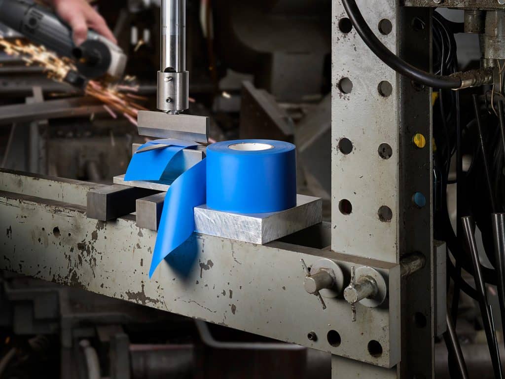 A roll of blue tape sits on a metal surface in an industrial manufacturing workshop, with machinery and tools visible. A person in the background uses a power tool, producing sparks. Various cables and bolts are attached to the metal structure.