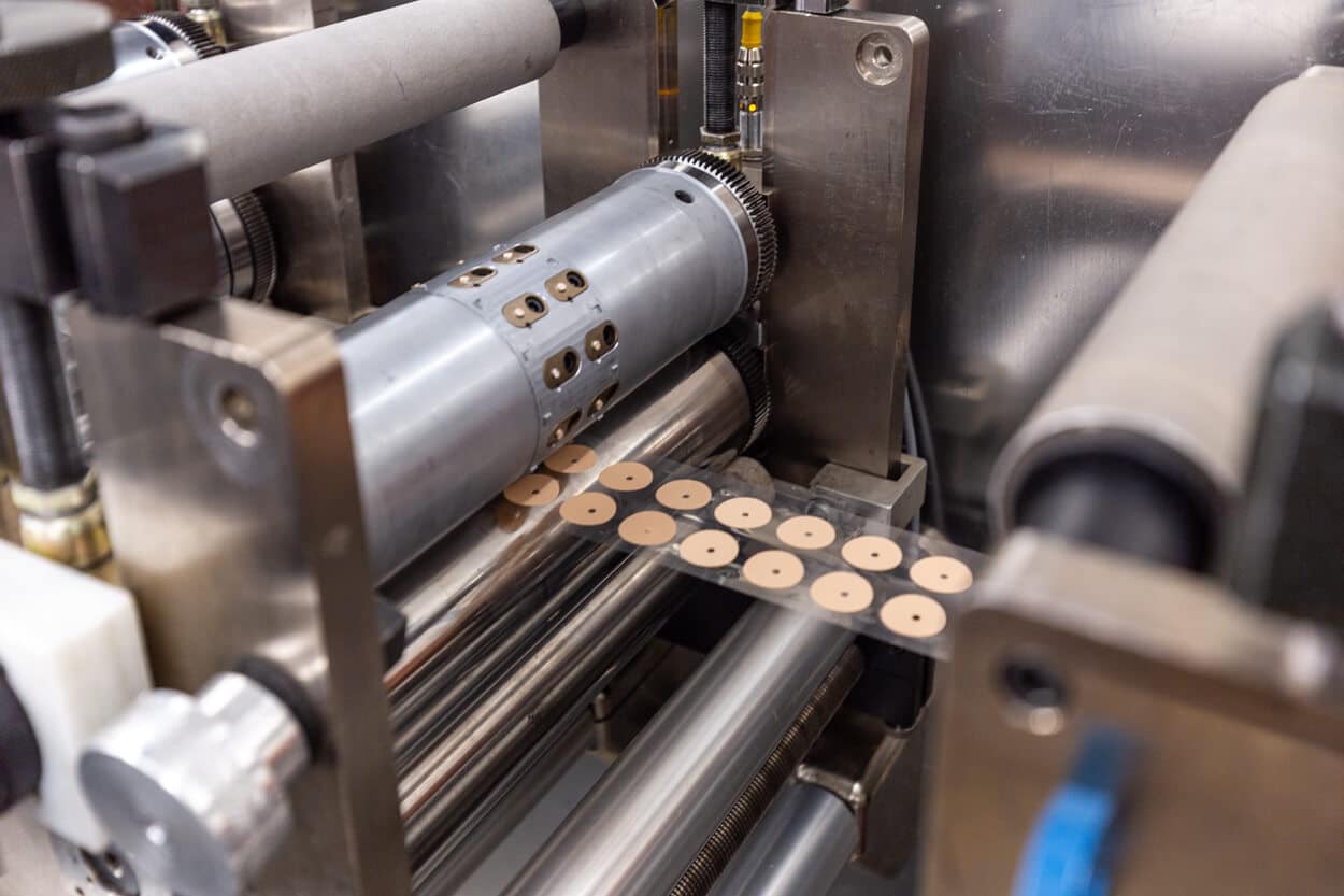 Close-up of an industrial machine with rollers slitting and cutting out small round brown disks from a strip of material, likely part of a contract converting or industrial manufacturing process.