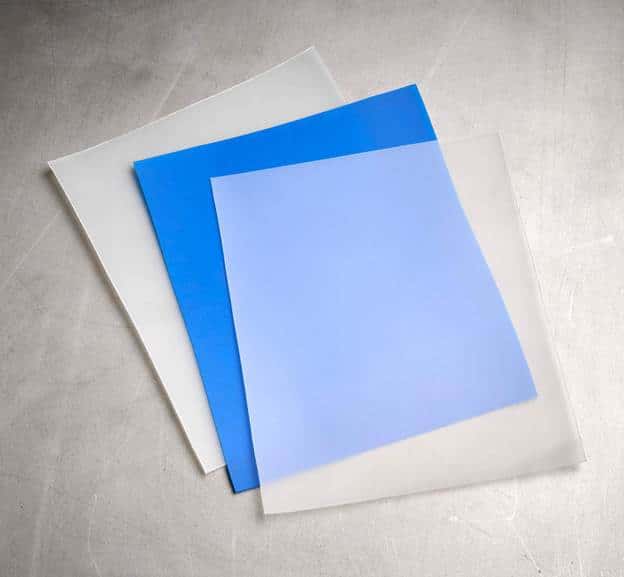 Three translucent sheets, one white, one blue, and one light blue, are stacked and slightly fanned out on a gray surface—perfect for slitting or other contract converting processes in industrial manufacturing.