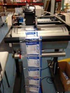 A machine for contract converting prints and cuts Essential packaging in an industrial manufacturing facility, with plastic packets feeding onto a conveyor belt. Boxes and equipment are visible in the background.