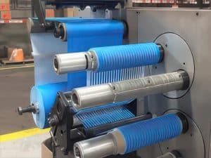 A close-up of an industrial machine with blue plastic film being unrolled and processed through several metal rollers in a factory setting, showcasing contract converting and slitting in industrial manufacturing.