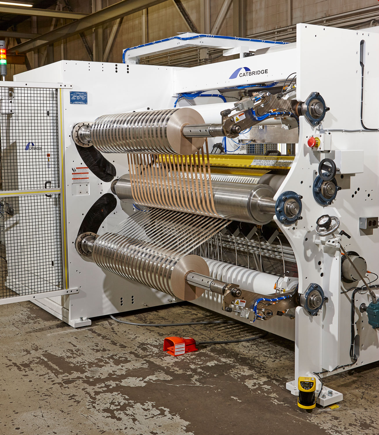 A large industrial slitter rewinder machine with multiple rollers is used for contract converting in industrial manufacturing, processing wide rolls of material into narrower strips. The white machine features metallic parts and side safety barriers.