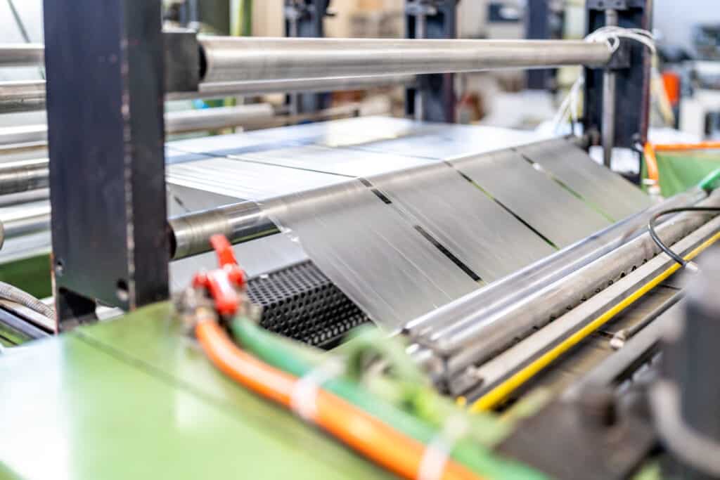Close-up of a machine slitting and cutting sheets of clear plastic film on rollers in an industrial manufacturing setting, with visible metal rods, orange wiring, and green machine parts.