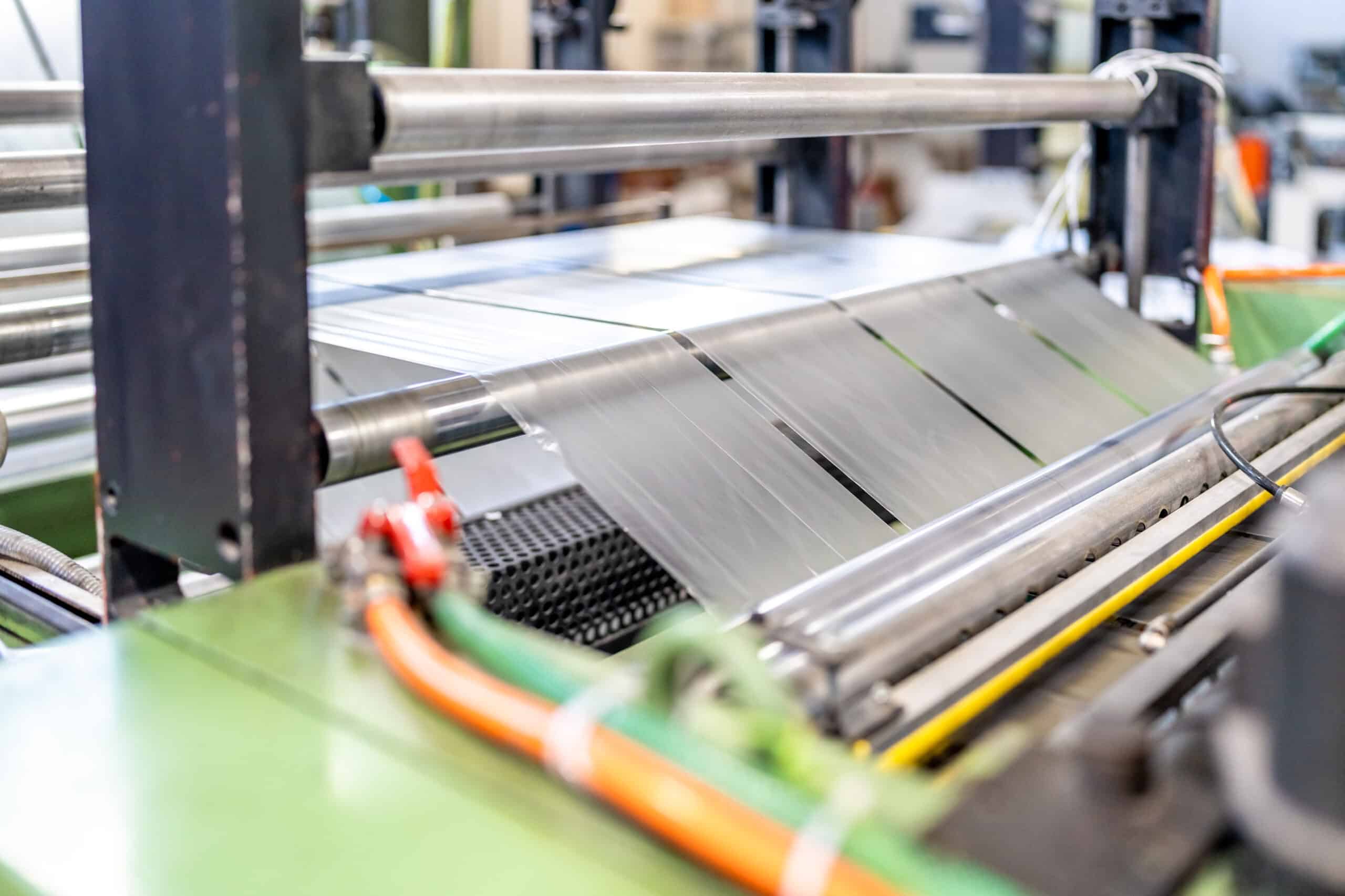 Close-up of a machine slitting and cutting sheets of clear plastic film on rollers in an industrial manufacturing setting, with visible metal rods, orange wiring, and green machine parts.
